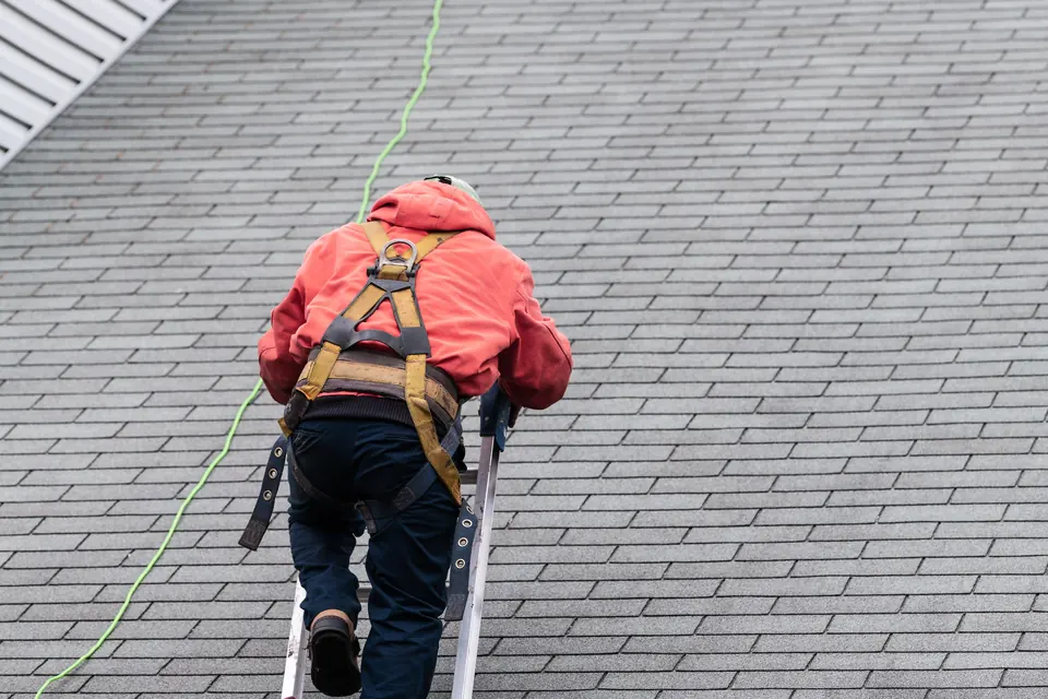 Roofer in a safety harness installing asphalt shingles on a pitched roof